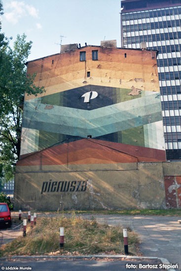 Ten kolorowy mural znajduje się na południowej ścianie kamienicy przy al. Politechniki 20. Jeśli by się obrócić o 180 stopni, zobaczylibyśmy jeszcze jeden mural (zobacz następny obiekt). Obie reklamy zamykają posesję 22/24 w wielobarwnym
